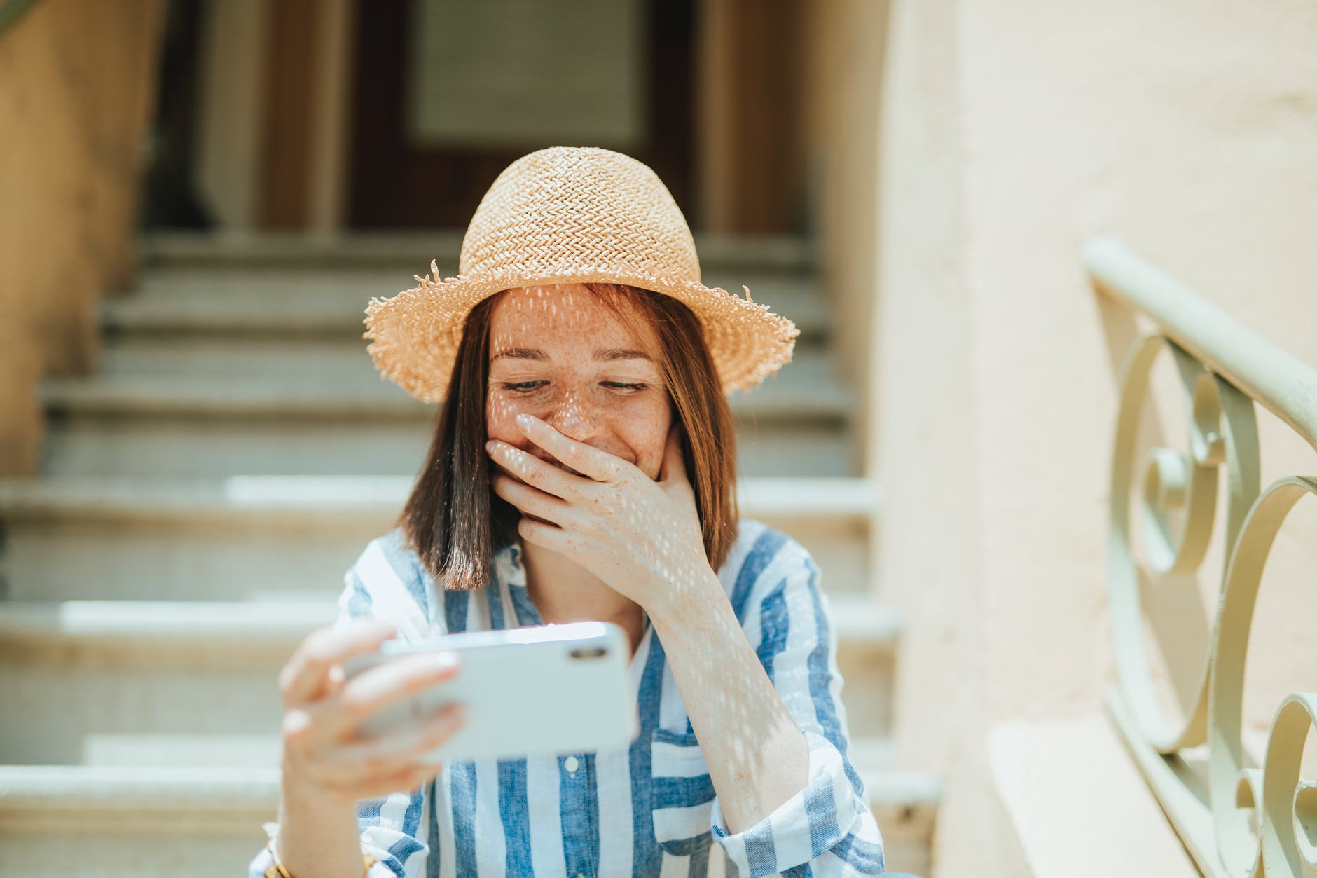 photo of woman smiling while siting on stairs and using white smartphone