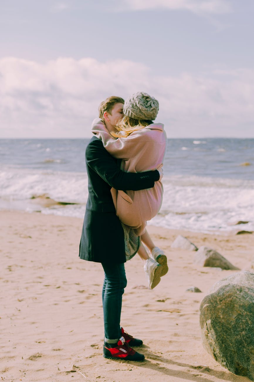man in black overcoat and blue denim jeans kissing while carrying a woman in pink overcoat and knit cap on shore at daytime